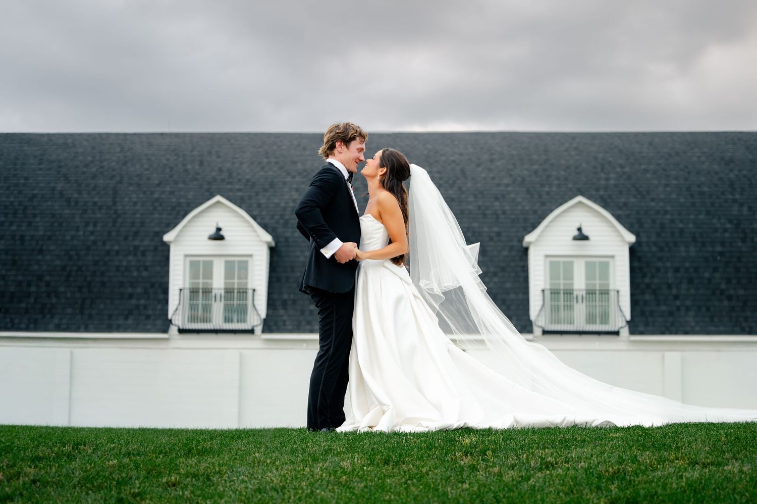 Bride and groom share a romantic kiss outdoors on green grass with a white building backdrop on their wedding day.