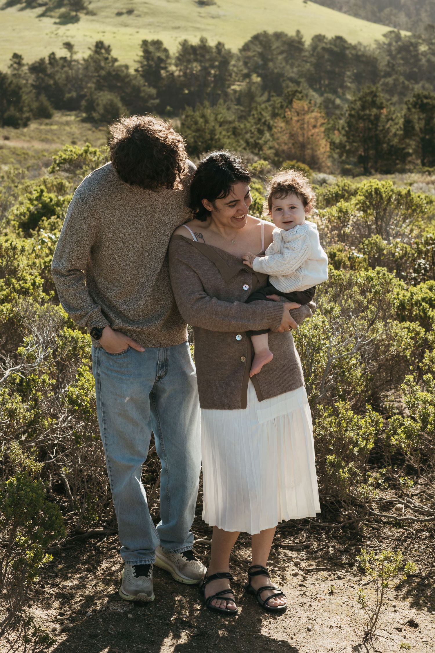 Family at beach with parents helping toddler walk on sandy shore, wearing cozy knit sweaters on a sunny day.