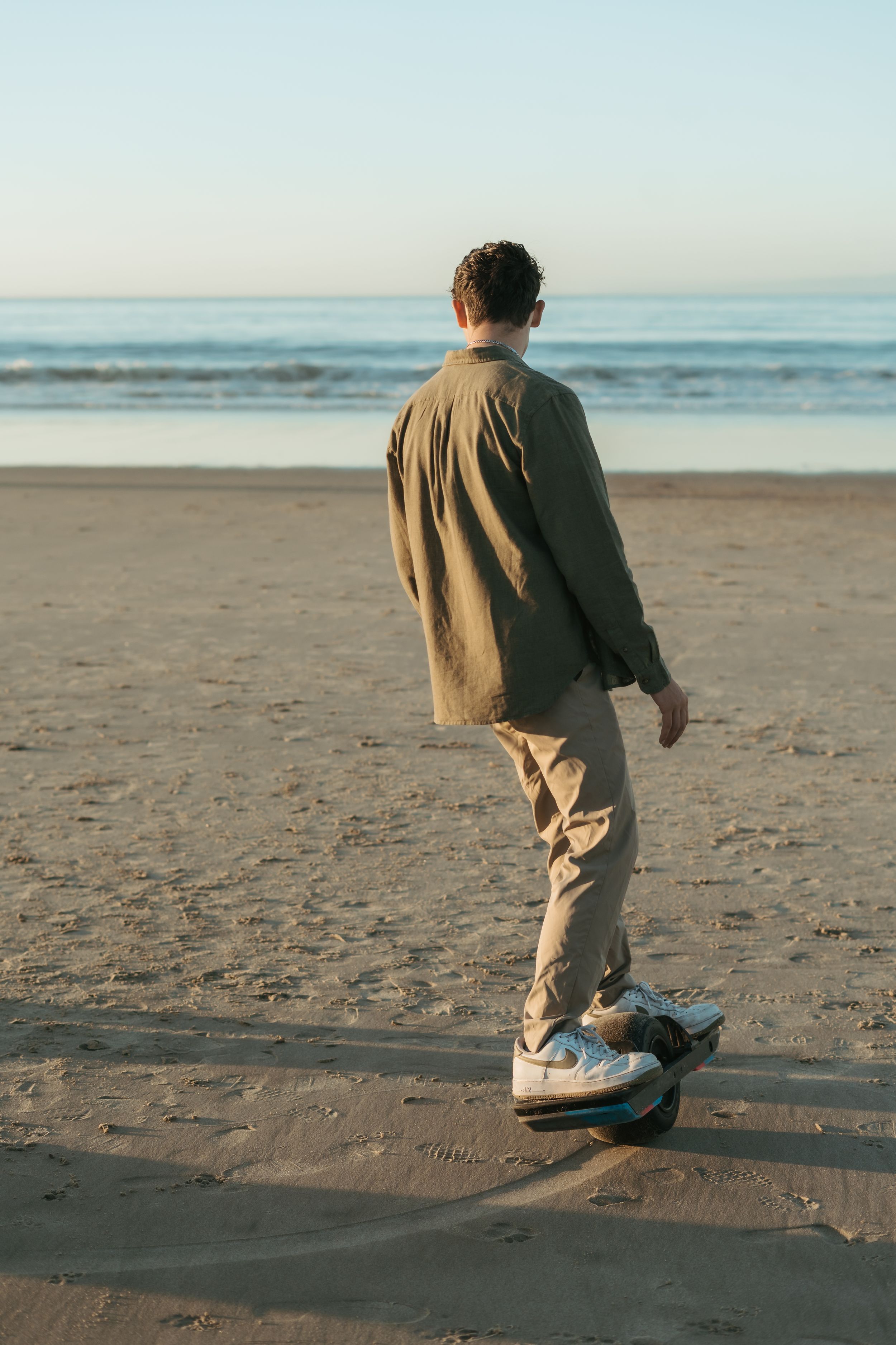 Man in olive jacket and khaki pants riding a skateboard on a sandy beach with ocean waves in the background.