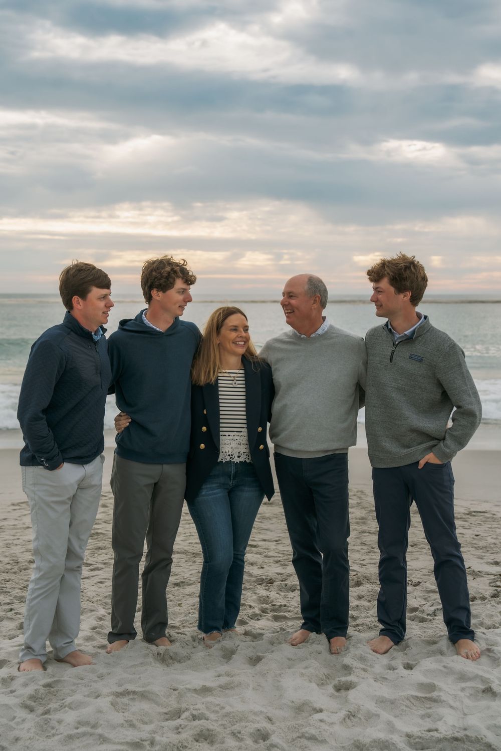 Family of five smiling together on a sandy beach at sunset, wearing casual coastal attire.