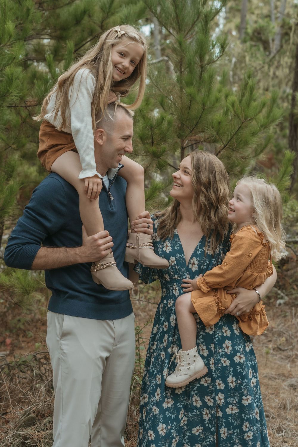 A happy family of four poses outdoors in a pine forest, with parents holding two young blonde girls.