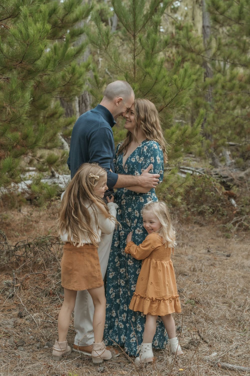 Family of four posing in pine forest, parents embracing while two young girls in earth-tone dresses hug them.