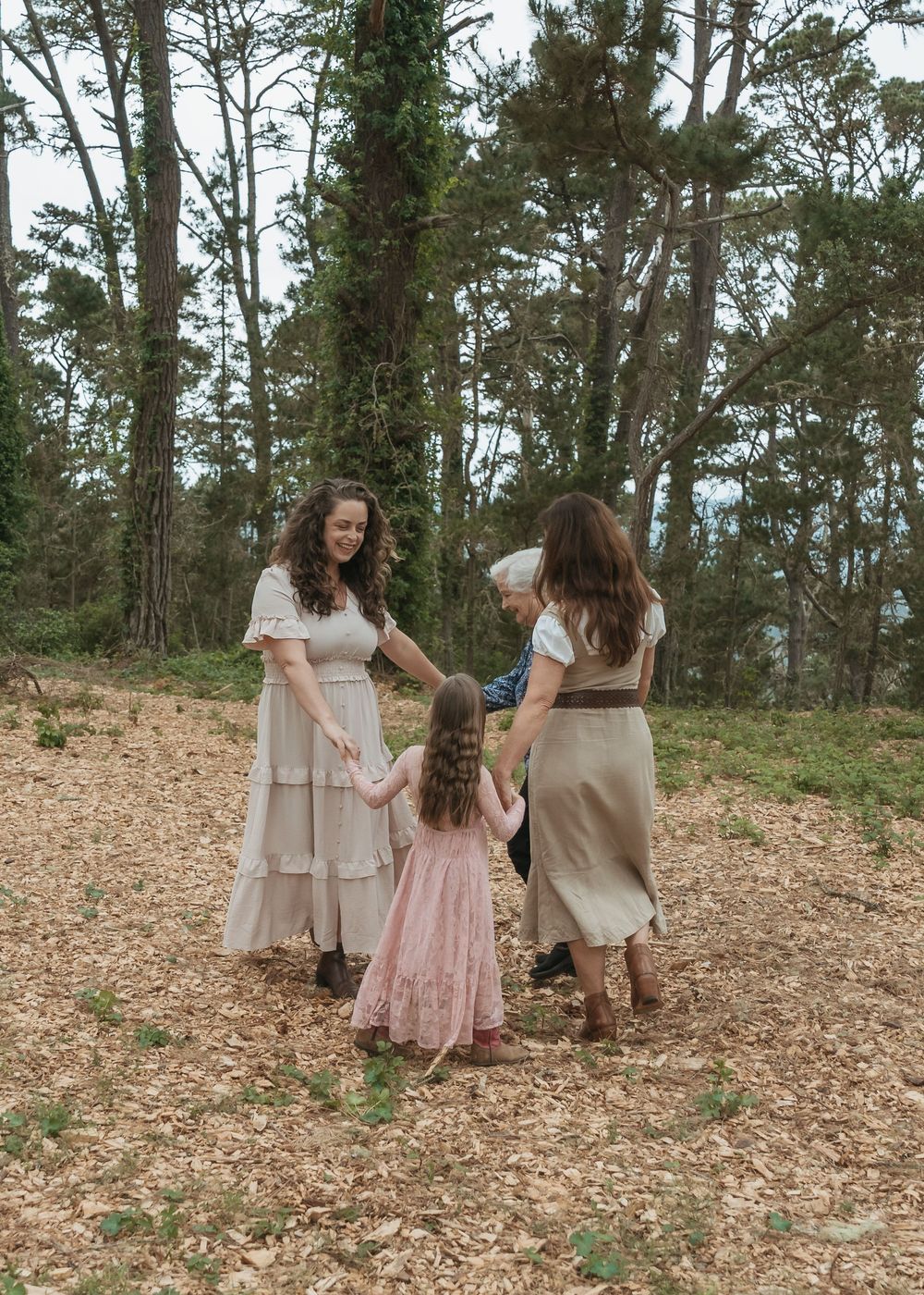 Two women and a young girl in flowing dresses hold hands in a circle on a woodland path surrounded by tall trees.