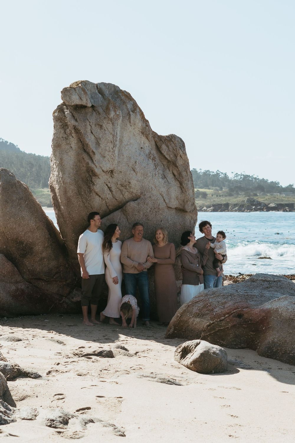 A family group poses together against large coastal rocks on a sandy beach with ocean waves in the background.