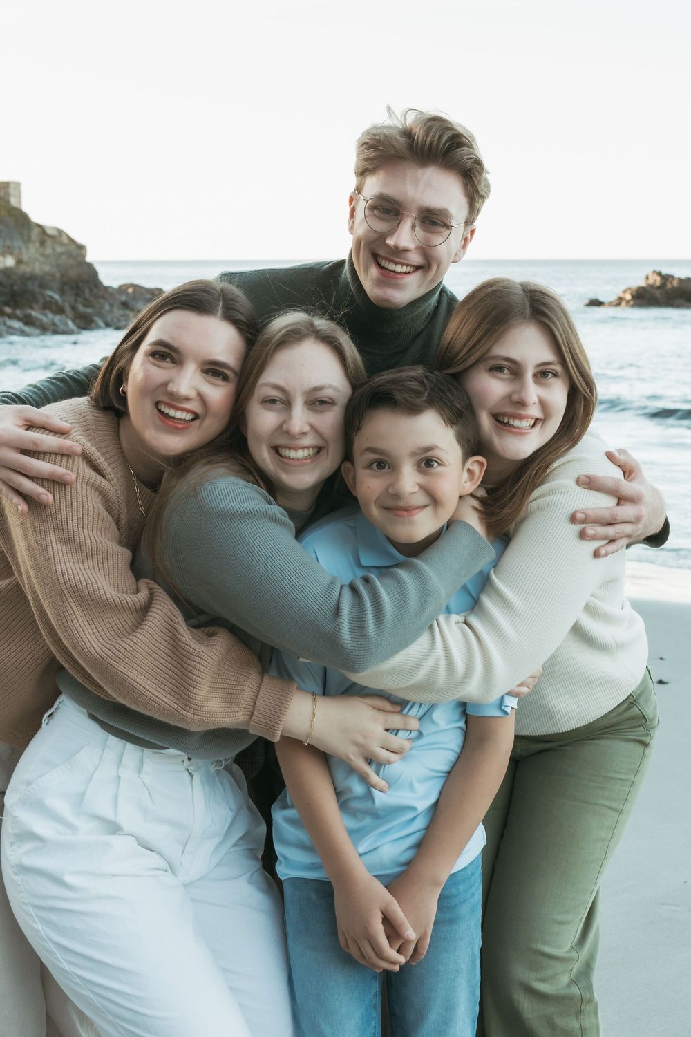 Happy family of five hugging together on a scenic ocean beach, smiling warmly in casual sweaters.