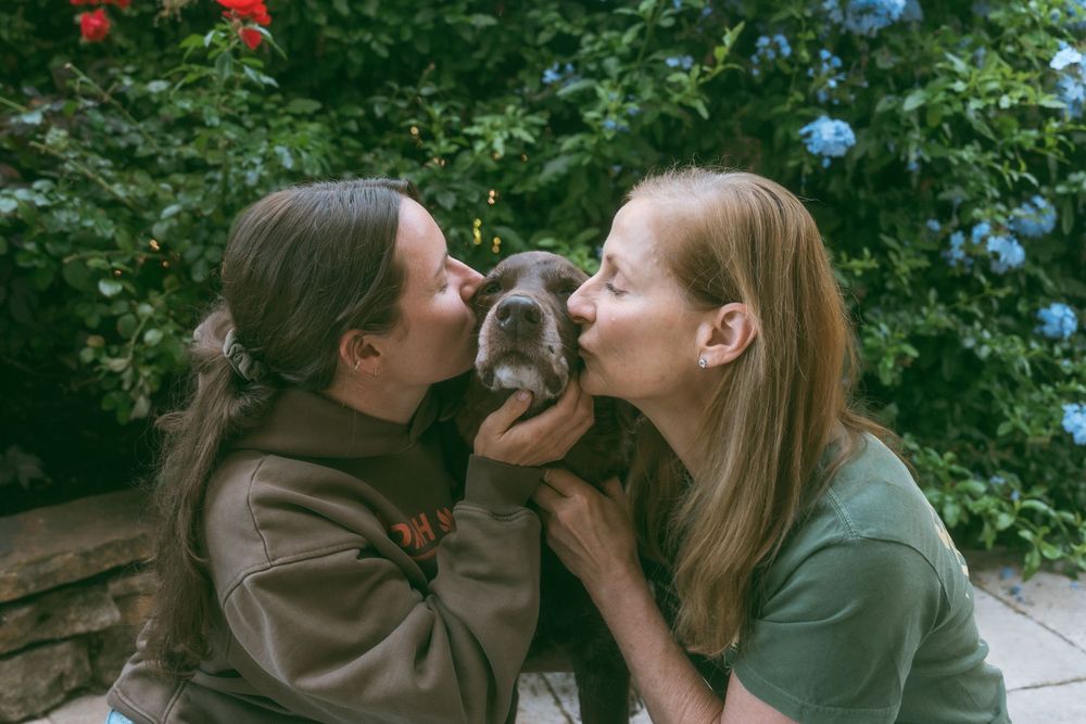 Two women kissing a cute gray puppy outdoors with lush green and blue flowering bushes in the background.