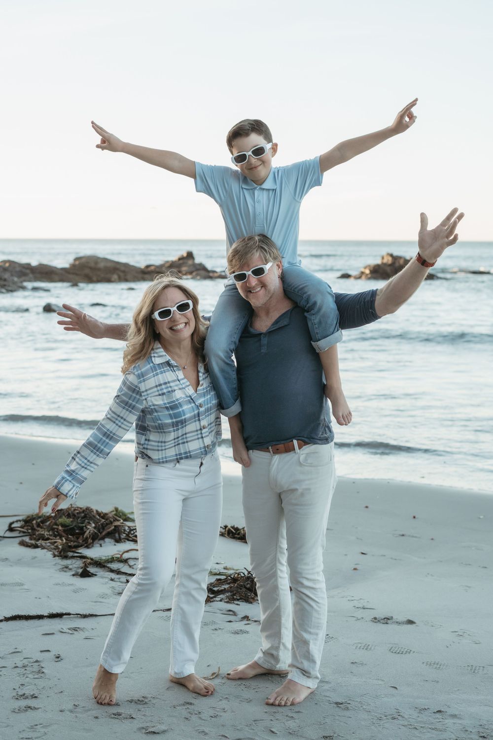 Happy family of three posing on a sandy beach, parents wearing sunglasses with child sitting on dad's shoulders, arms outstretched.