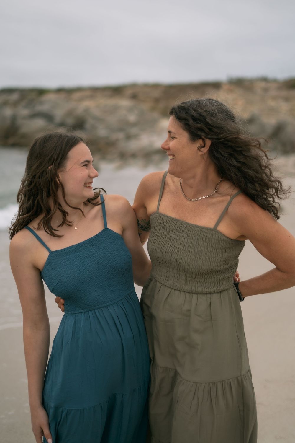 Two women in summer dresses smiling at each other on a beach, one in teal and one in olive green.