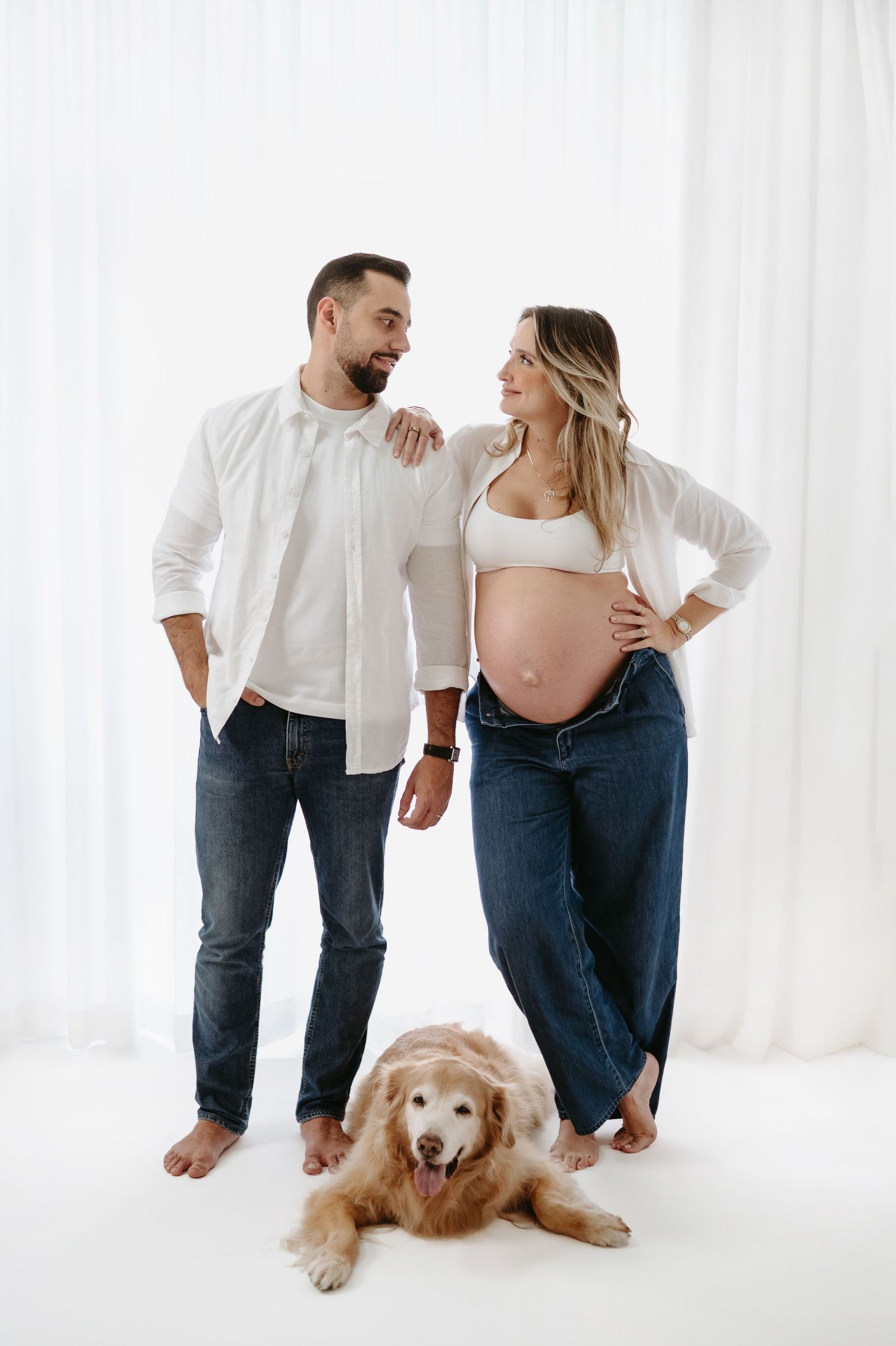 Pregnant couple in white and denim smiling at each other with a golden retriever dog lying at their feet.