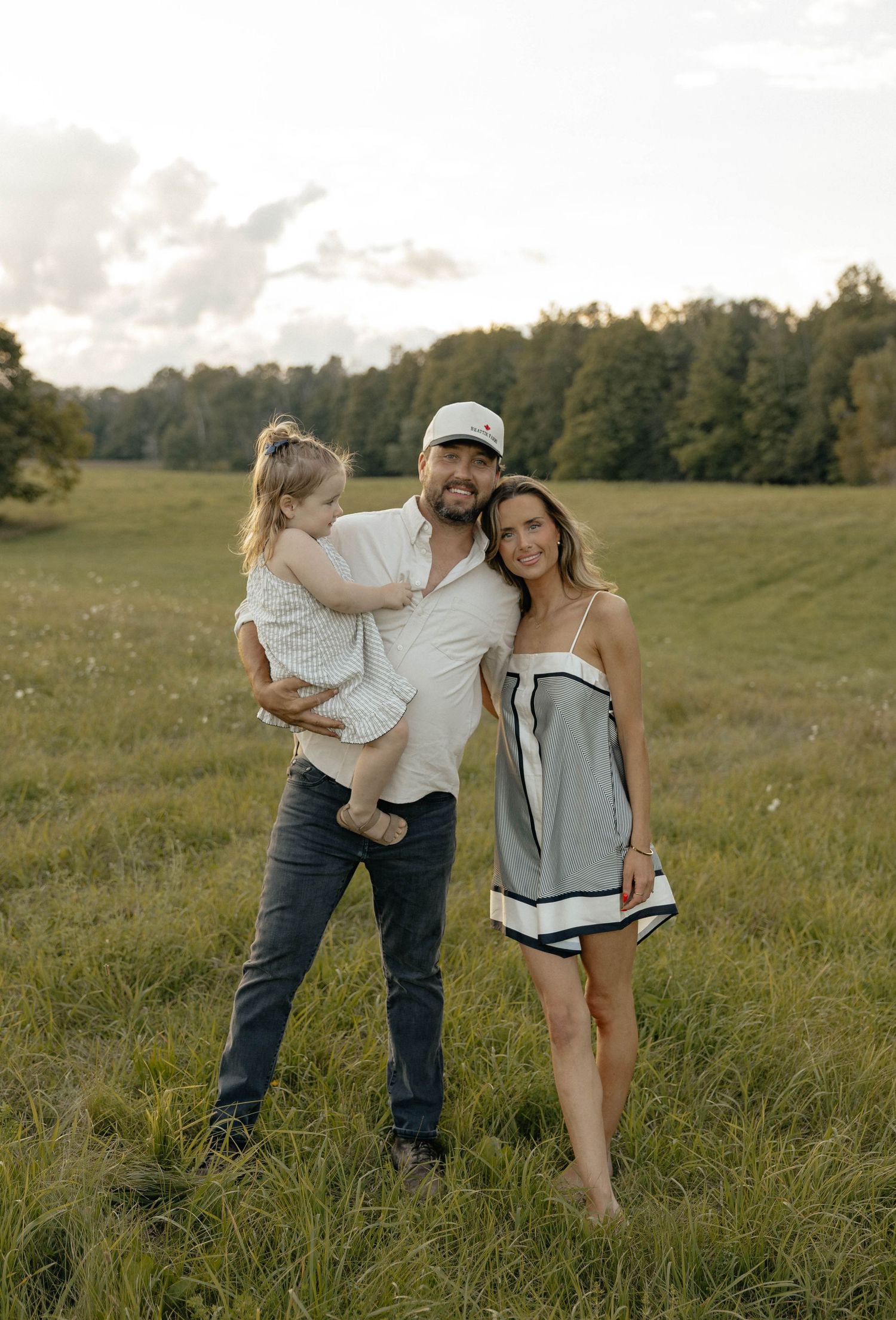 A happy family of three posing in a lush green meadow at golden hour, man holding young child beside smiling woman.