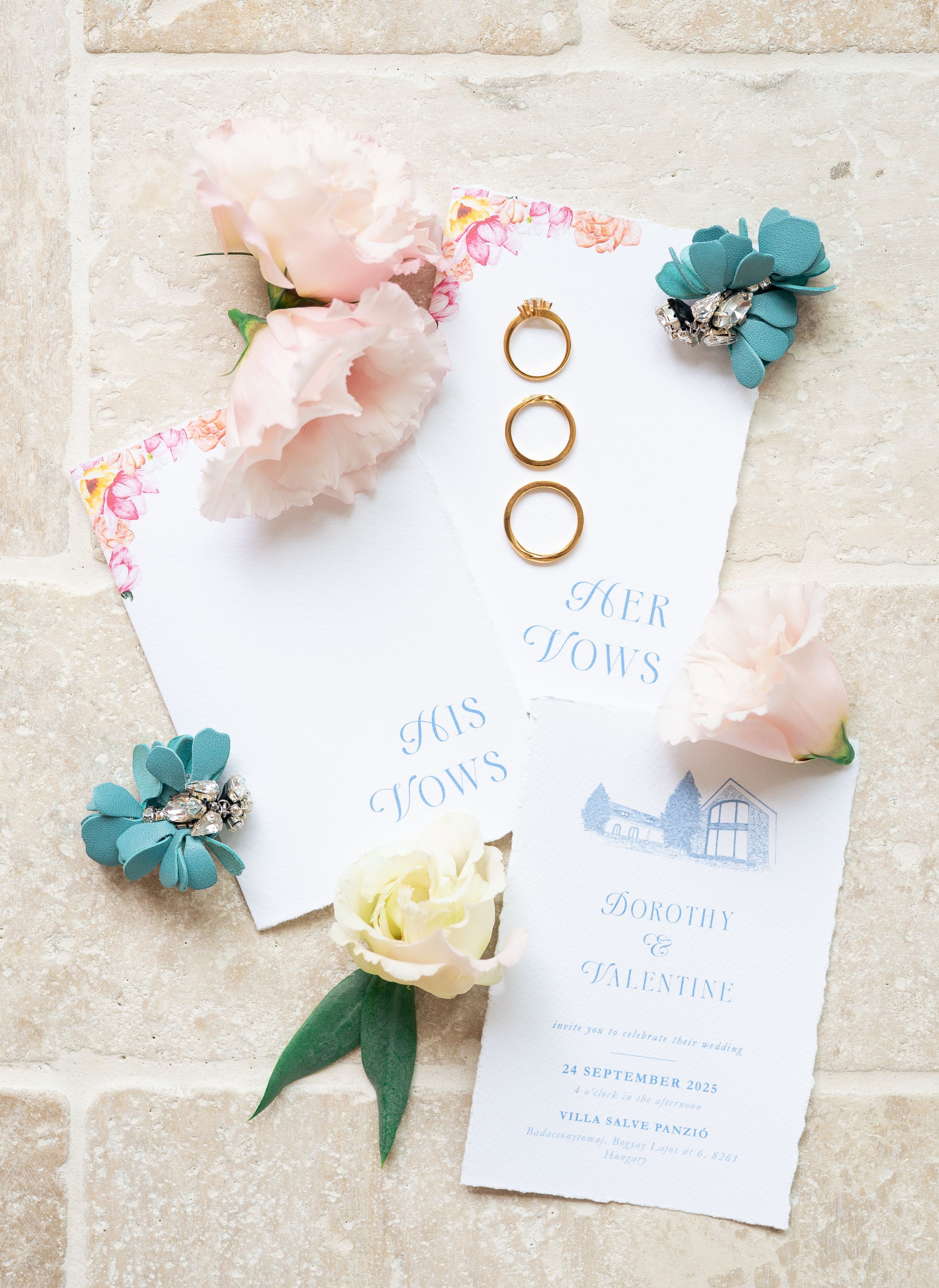 Wedding stationery flat lay with gold rings, vow books, floral invitation suite, and pink and teal flowers on stone.