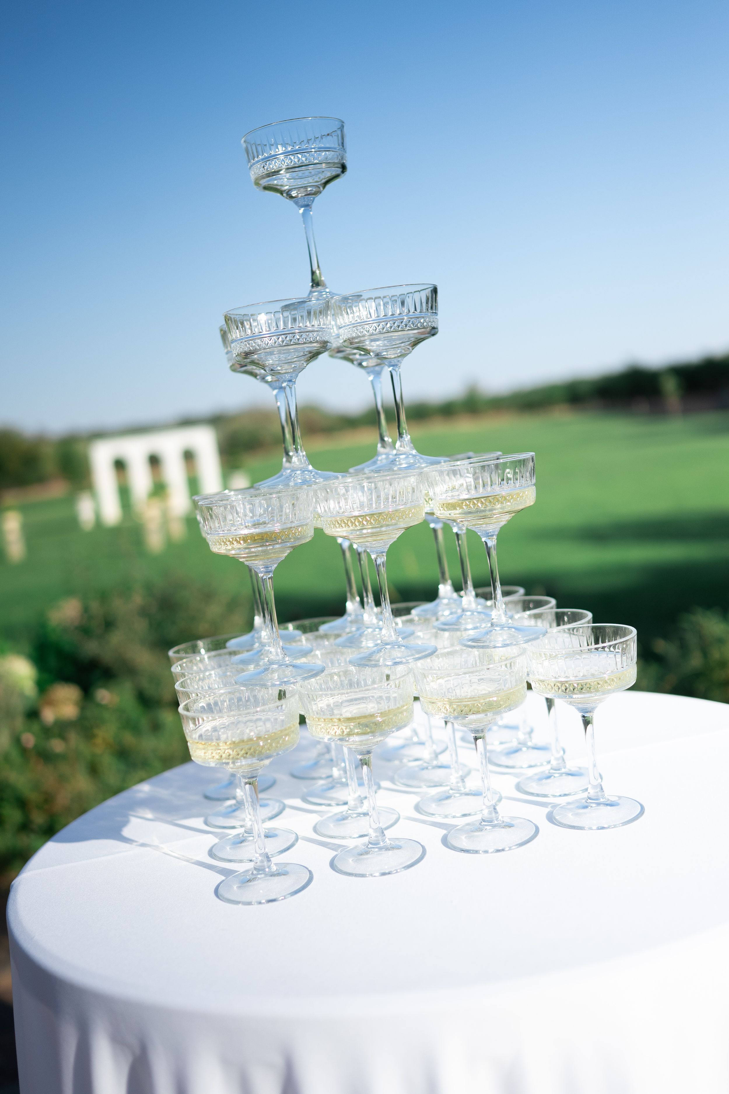 Crystal champagne glasses stacked in a pyramid tower at an outdoor wedding reception with green lawn background.