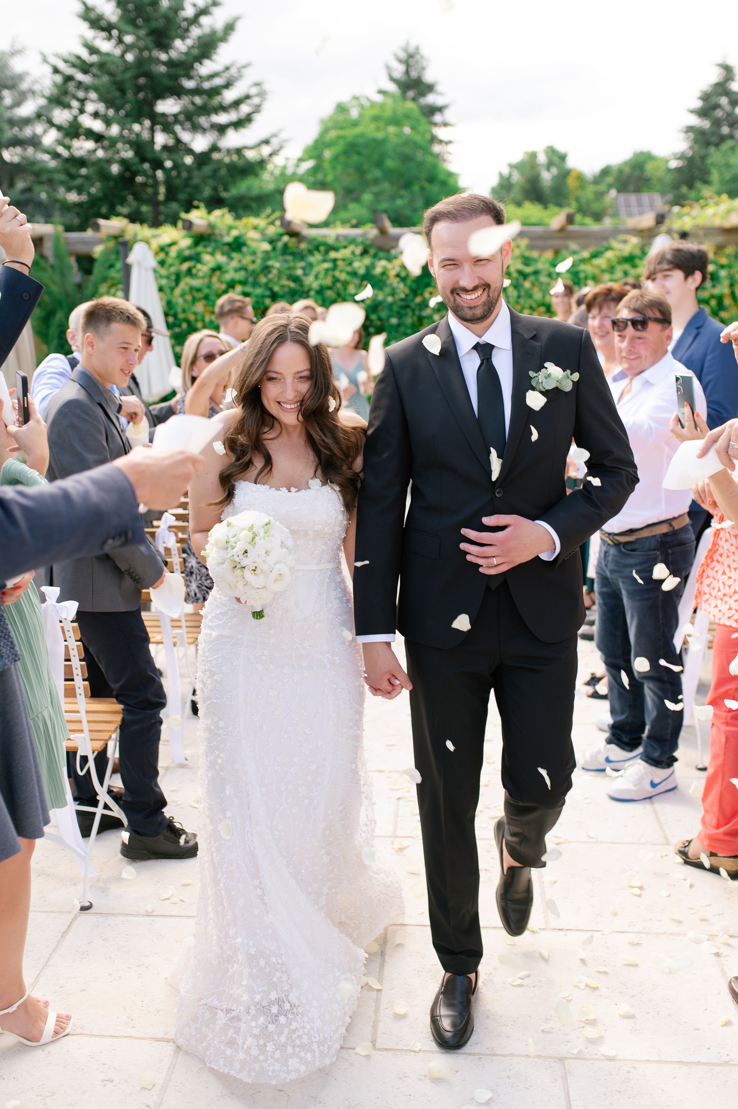 Joyful newlyweds walk down outdoor aisle as guests toss white flower petals in sunny wedding ceremony.