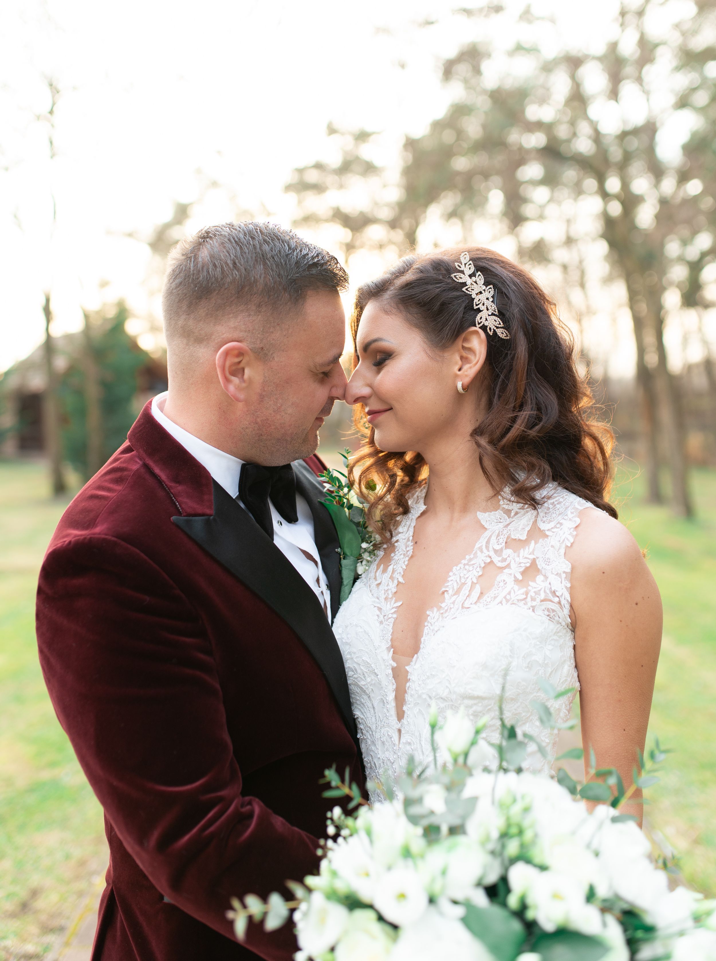 Bride and groom touching foreheads outdoors, groom in burgundy velvet tuxedo, bride in lace wedding gown holding white bouquet.