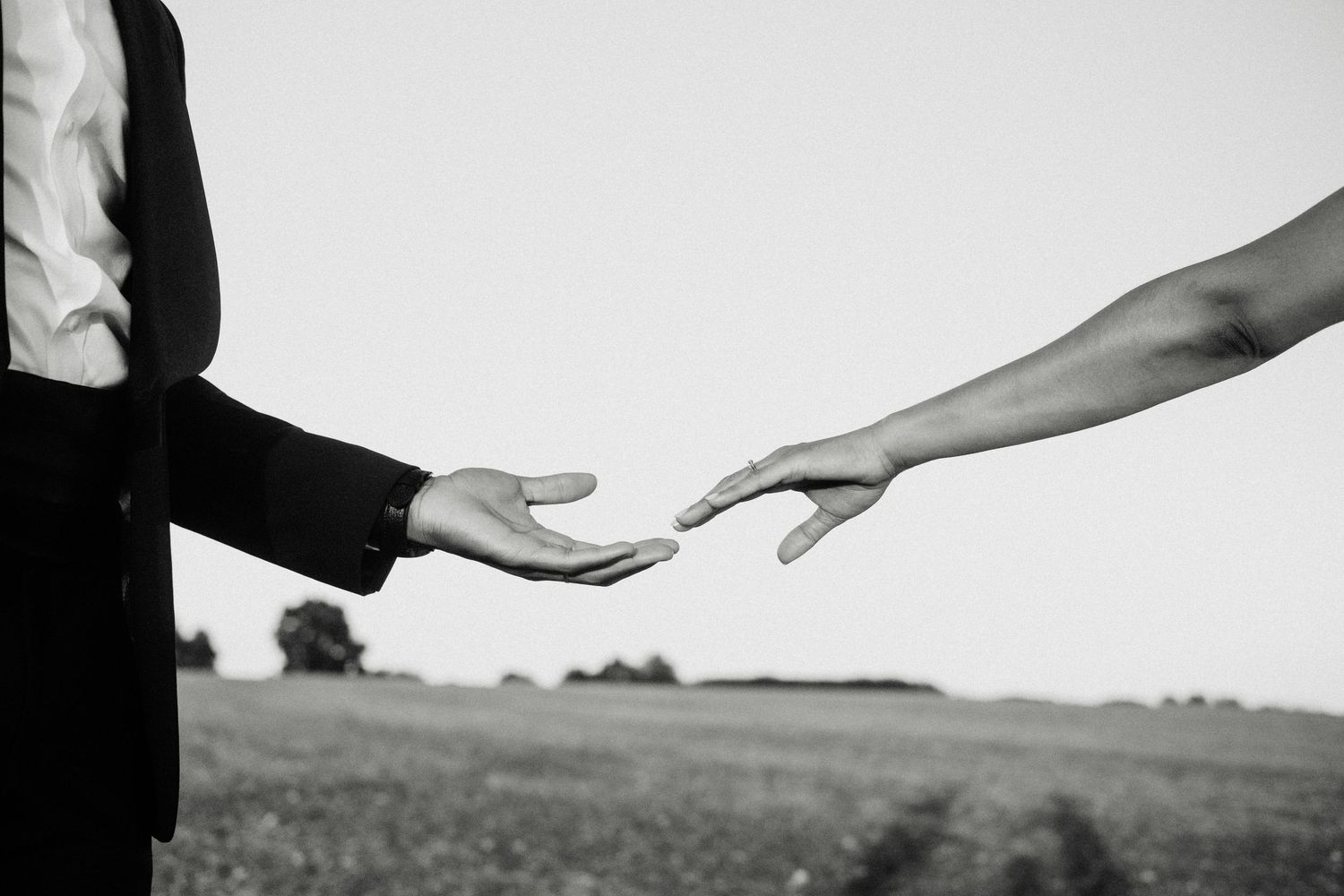 Black and white photo of two hands reaching toward each other over a field, evoking connection and romance.