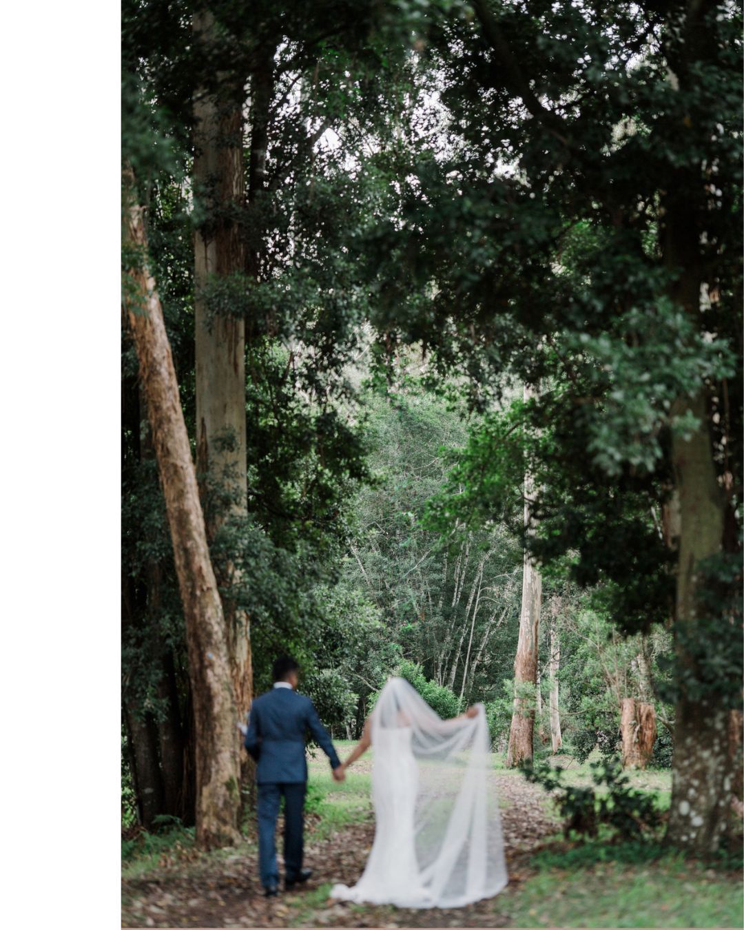 A bride and groom walk hand-in-hand down a lush forest path, her veil trailing gracefully behind them.