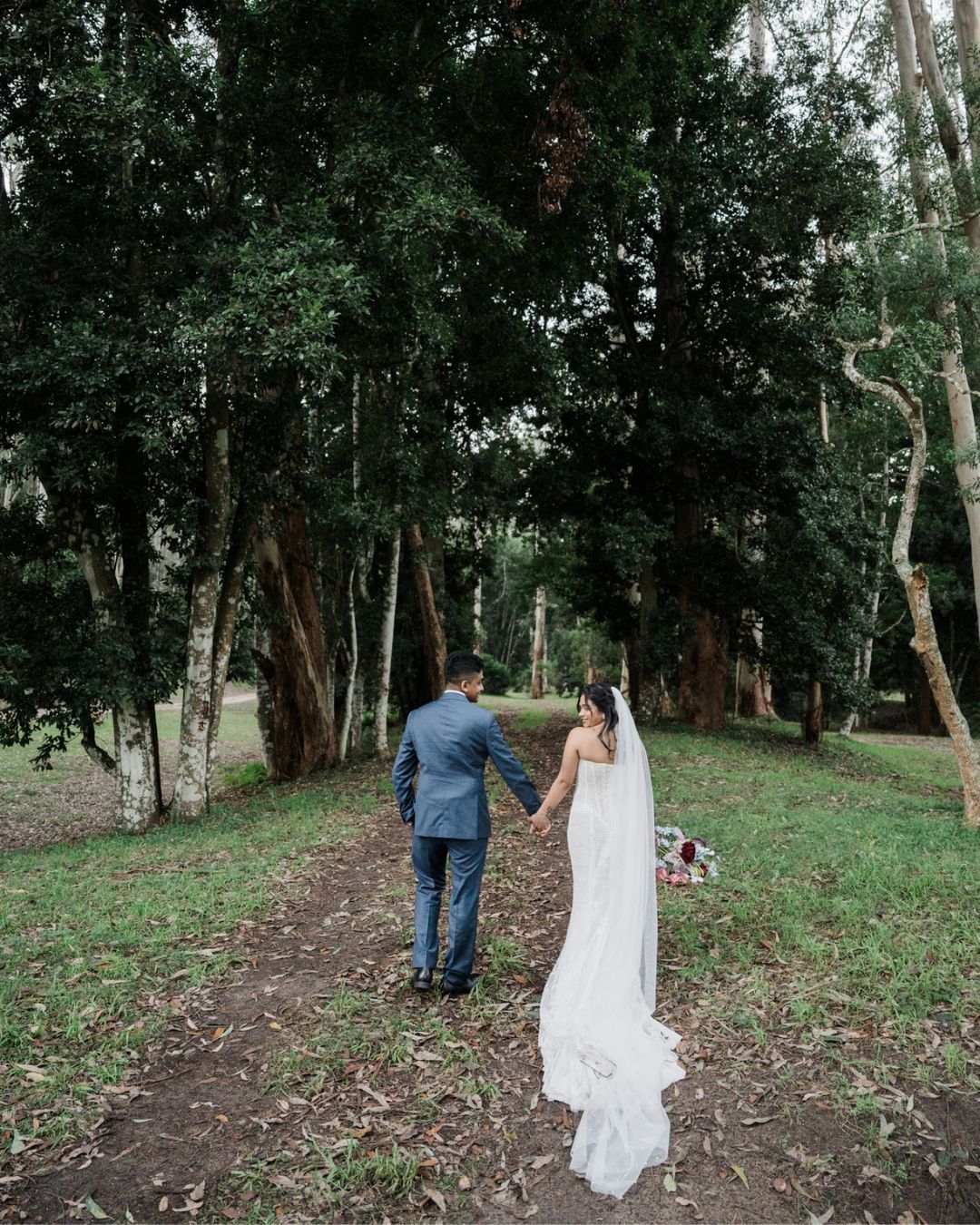 A bride and groom hold hands walking toward a lush forest on their wedding day.