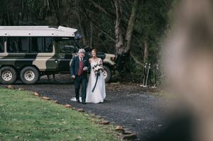 Bride walks with escort down gravel path toward wedding ceremony with vintage Land Rover in background.