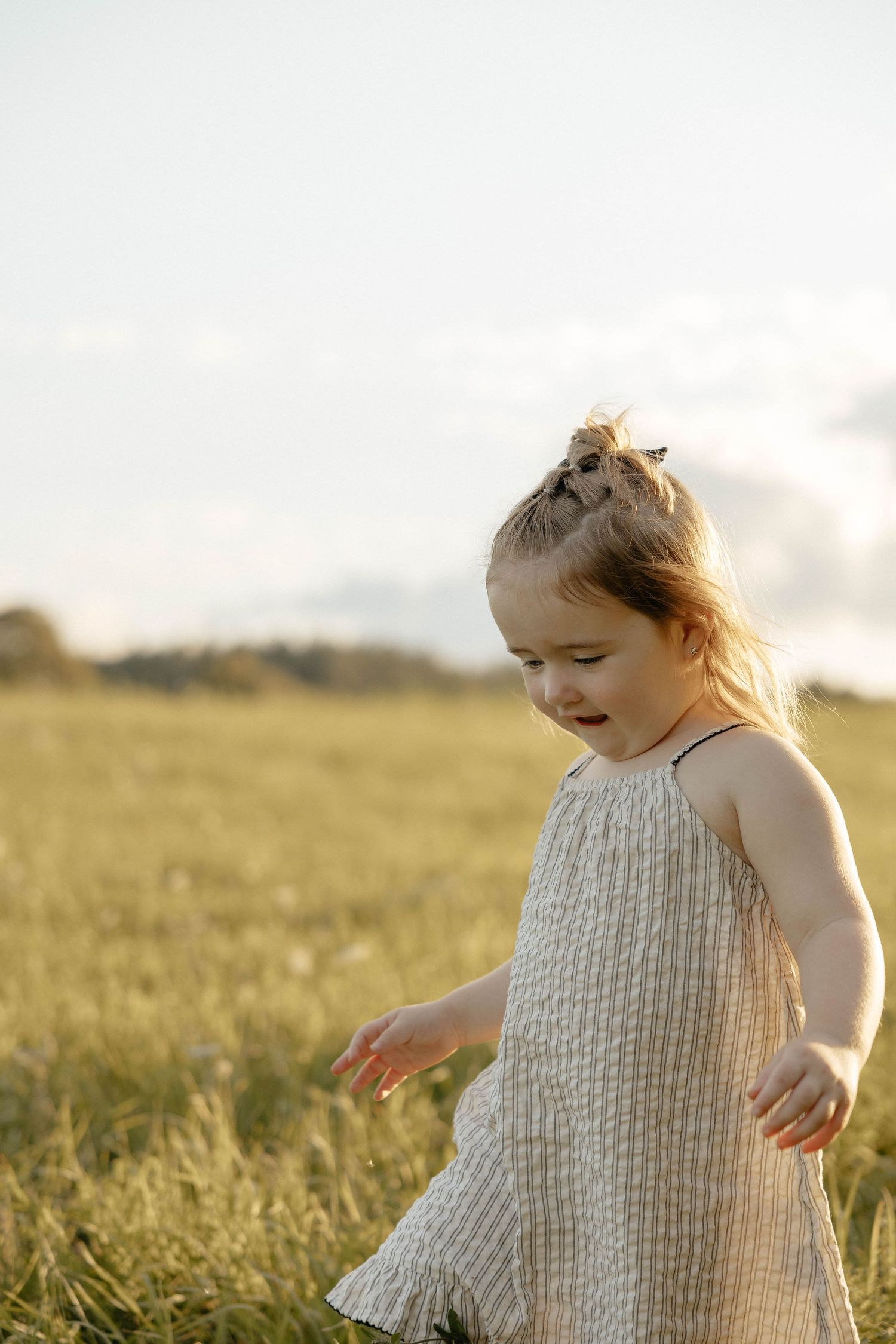 A young girl in a linen dress plays in a sunlit green field, smiling and enjoying the outdoors.