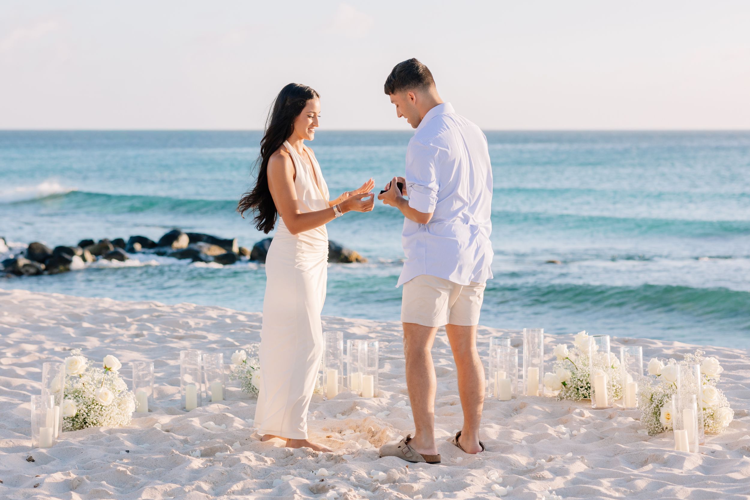 Couple exchanging rings during a romantic beach proposal surrounded by white floral arrangements and candles at sunset.