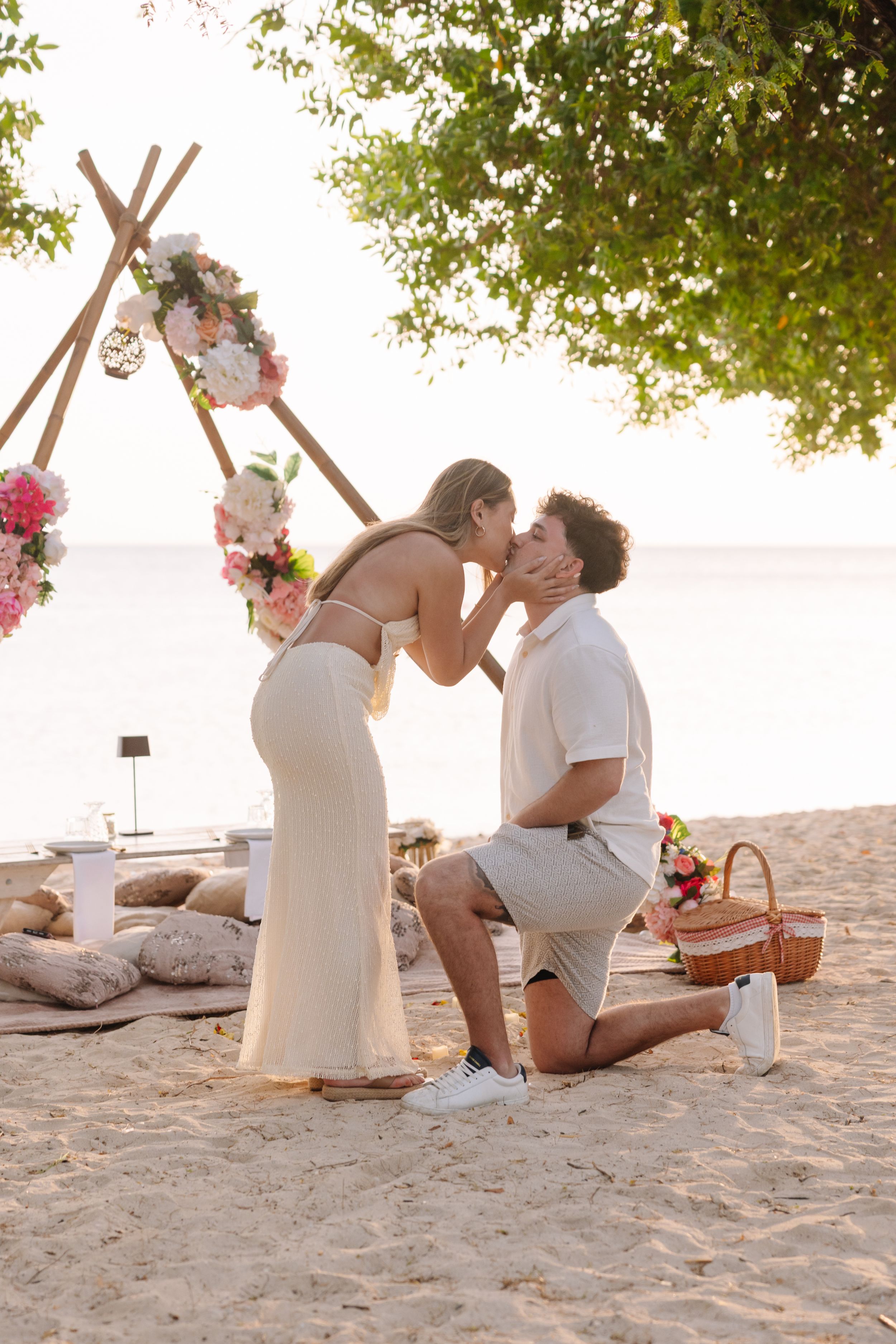 Man proposes to woman on beach with floral arch backdrop at sunset, romantic seaside engagement moment.