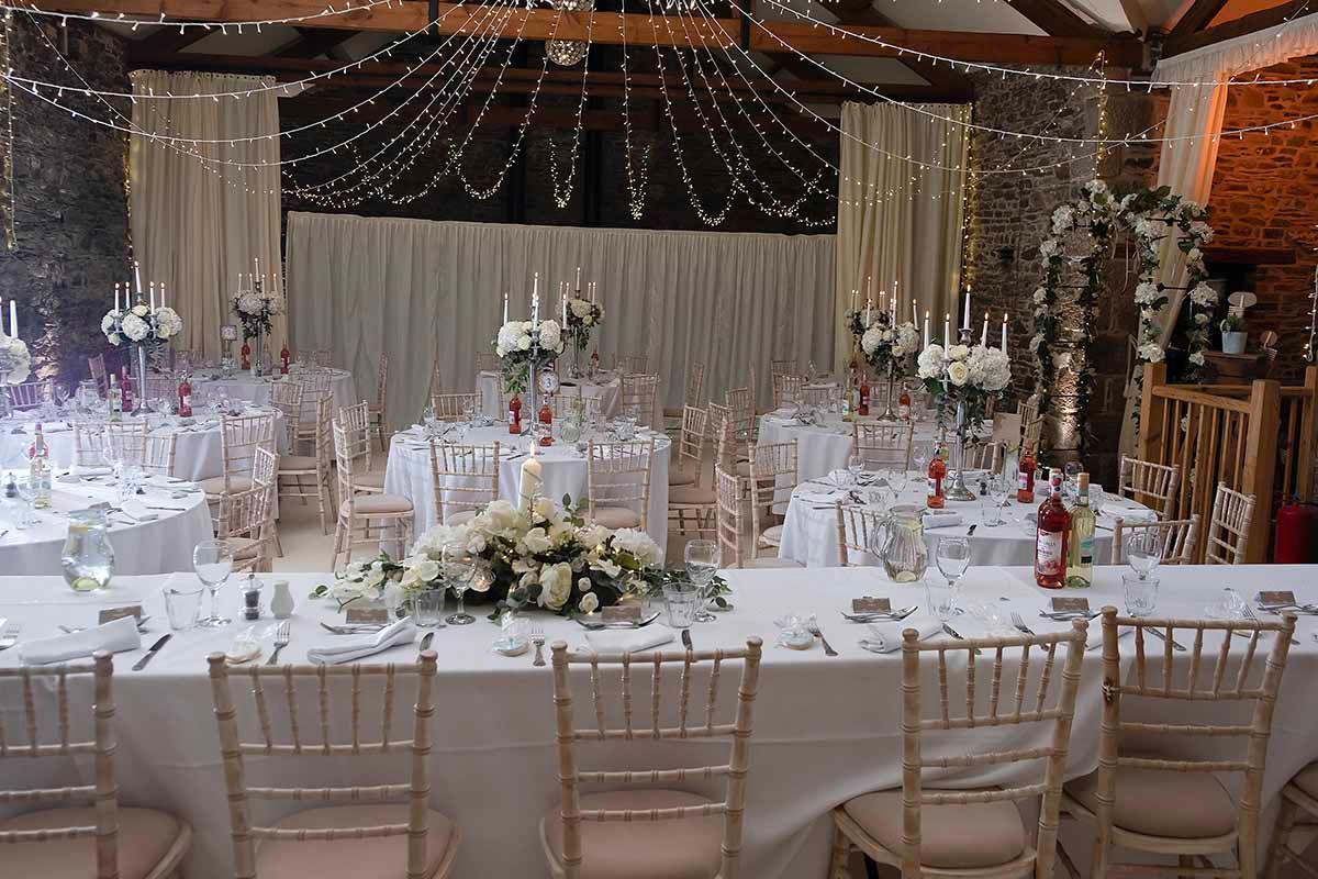Elegant wedding reception in a barn venue with fairy lights, white floral centre pieces, and gold chivalry chairs at New House Farm