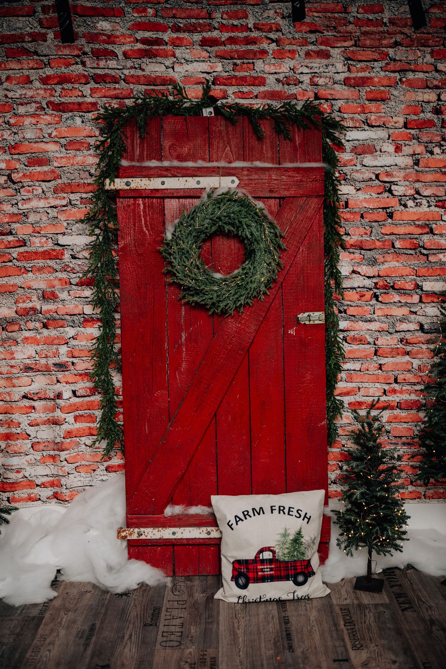 A festive green Christmas wreath with fairy lights and snow hangs on a rustic red wooden barn door with brick wall.
