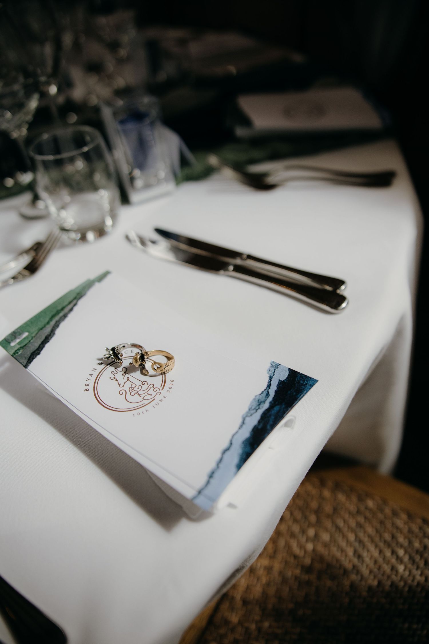 Wedding rings resting on a white menu card at an elegantly set reception table with silverware and glassware.
