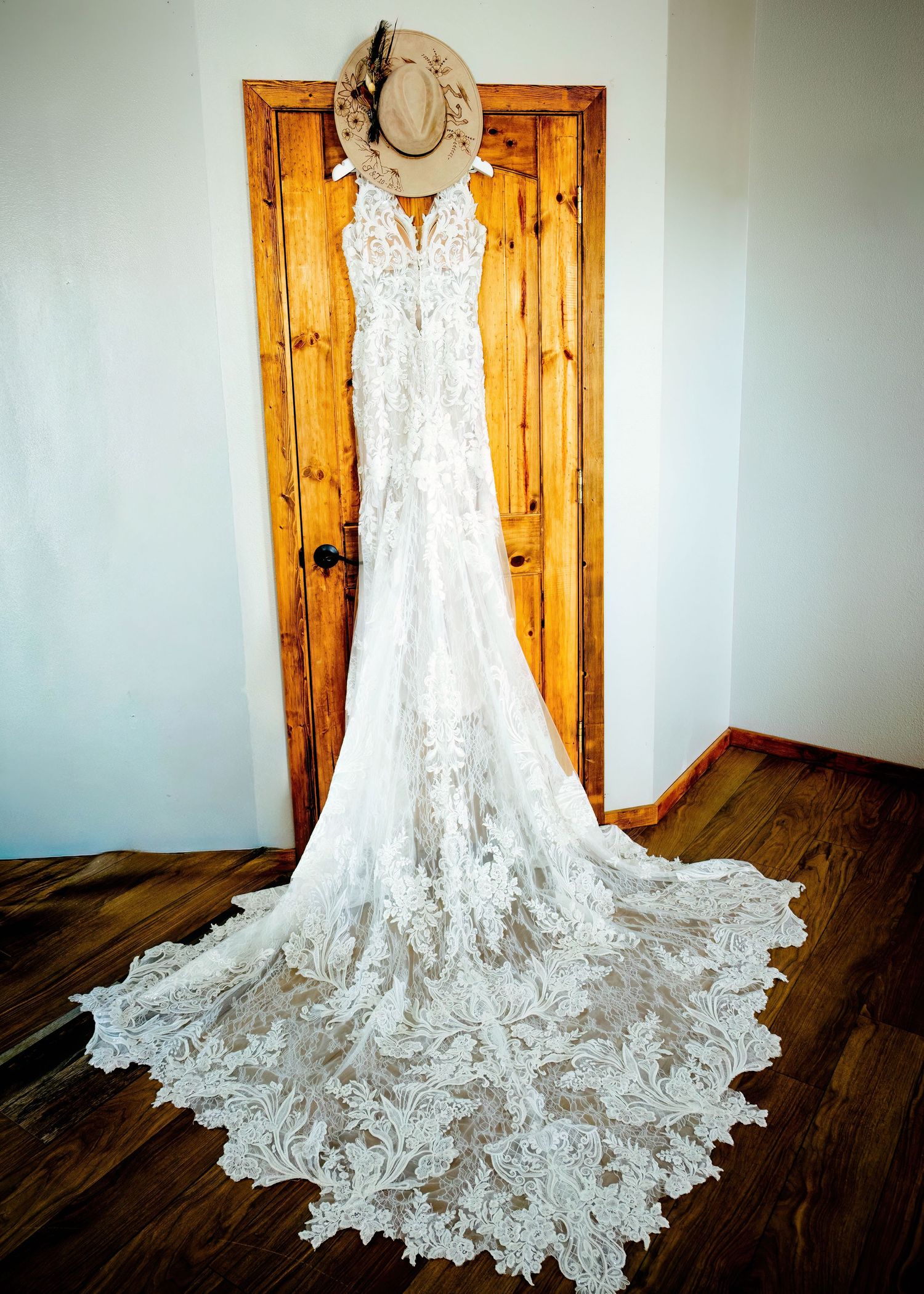 Elegant lace wedding dress with a long train hanging on a rustic wooden door, topped with a tan cowboy hat.
