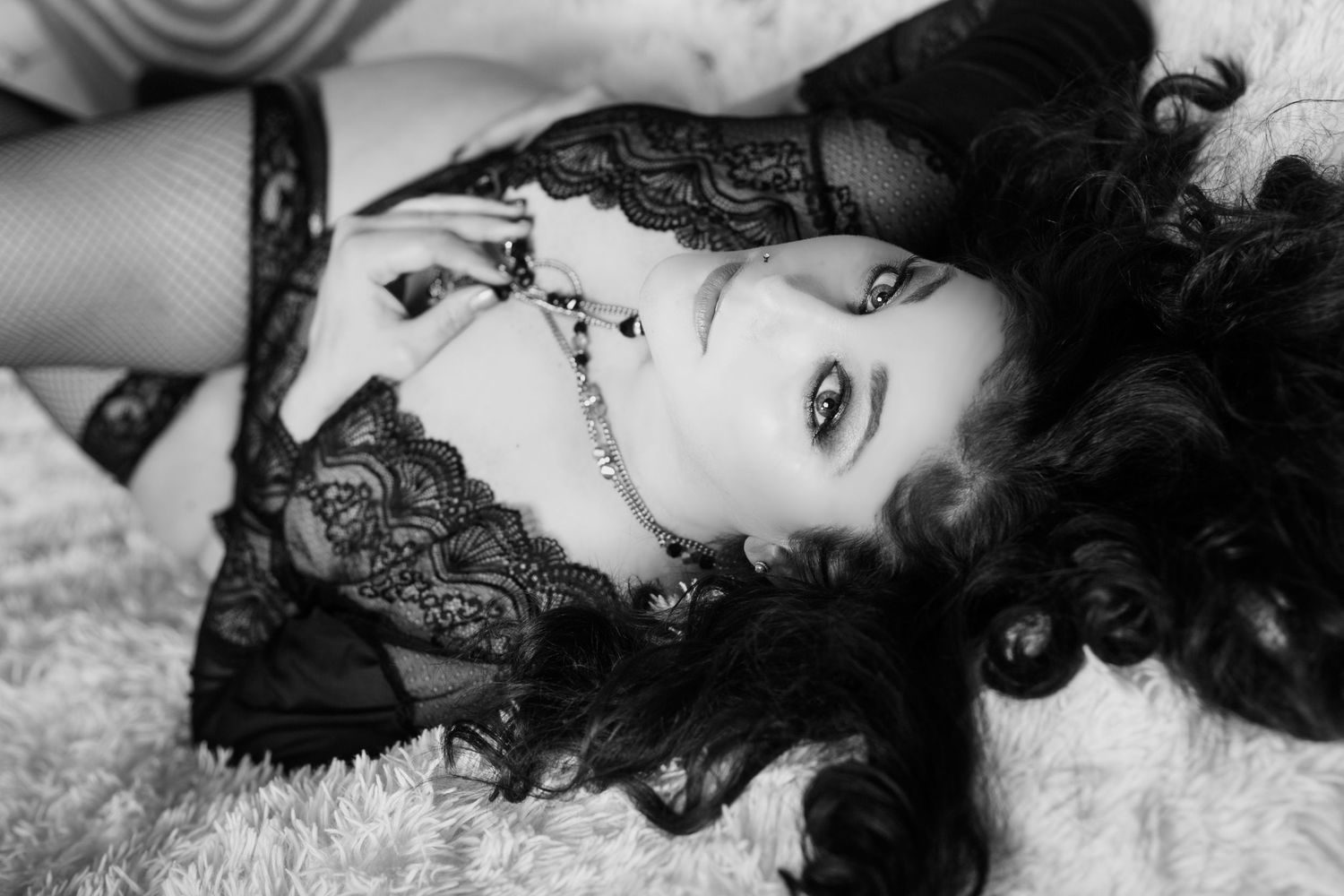 Black and white boudoir photo of a woman in lace lingerie lying on a fur rug with dramatic makeup.