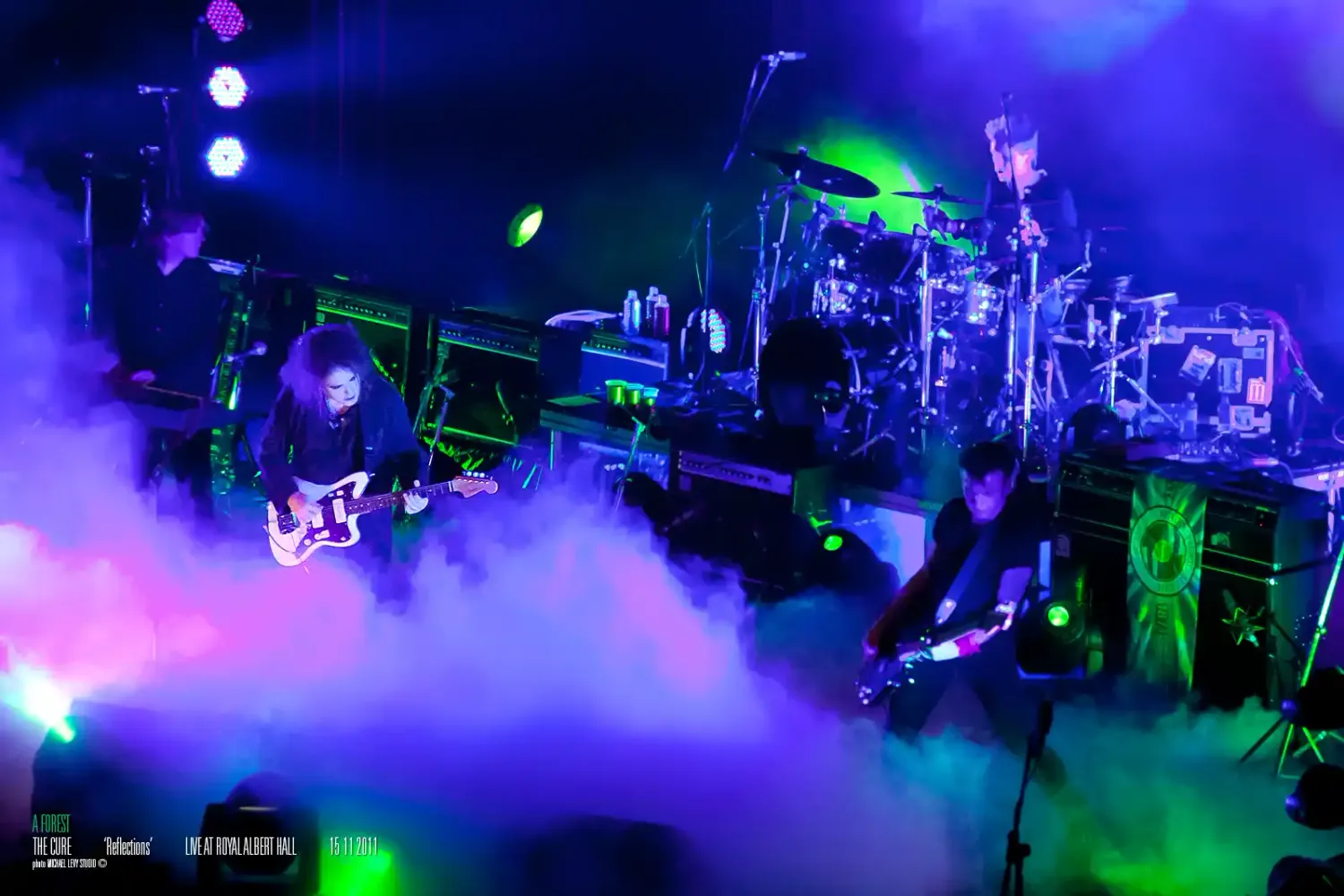 Rock band performs on fog-filled stage with vibrant purple and green lights illuminating musicians and equipment.