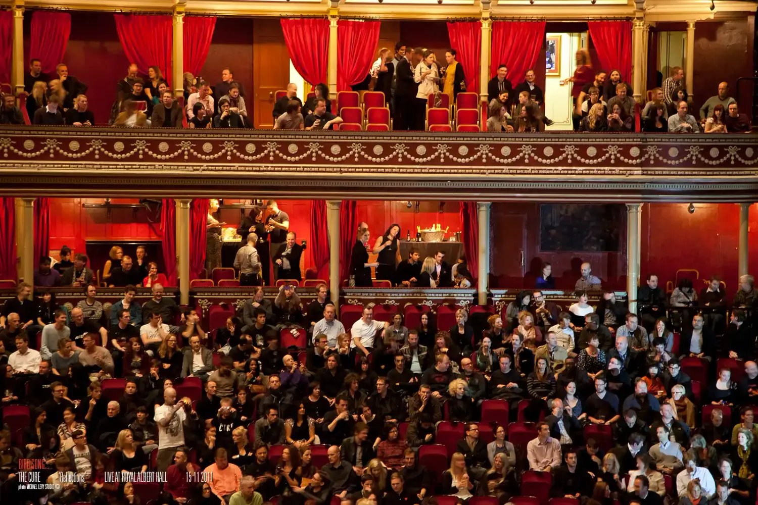 Elegant theater interior filled with audience members seated across multiple ornate balcony levels with red curtains.