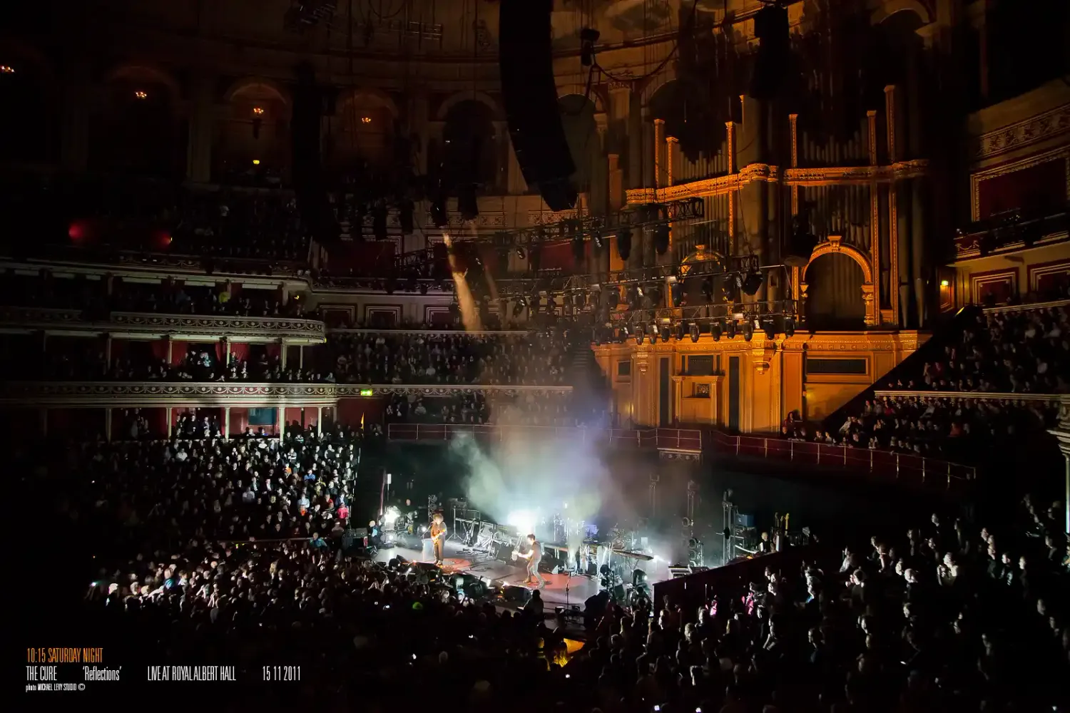 A packed concert hall with dramatic stage lighting illuminating performers on stage at a grand ornate venue.