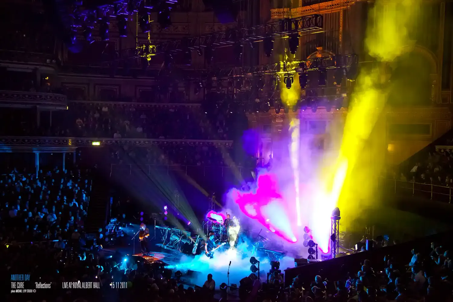 Vibrant concert stage with colorful pink, yellow, and blue lights illuminating performers at a packed indoor venue.