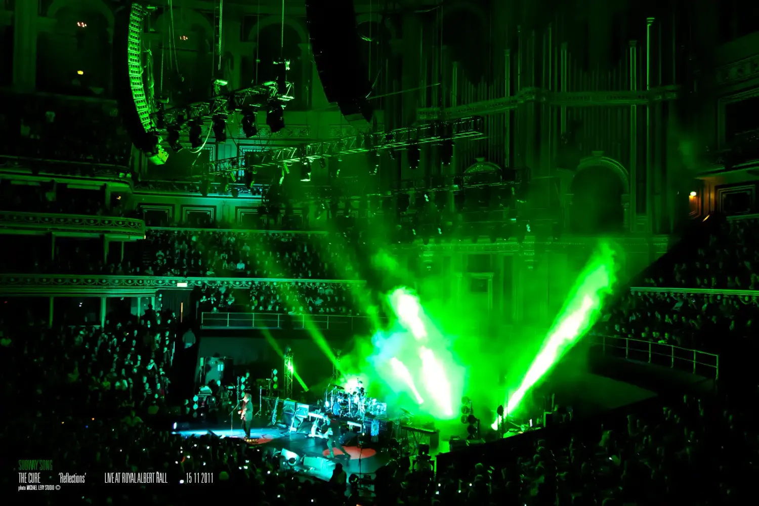 A packed concert venue bathed in vibrant green laser lights illuminating the stage and crowd from above.