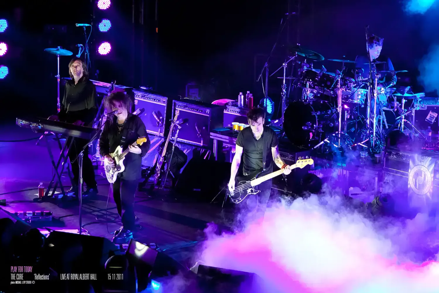 Rock band performing on a colorful stage with purple and blue lights, fog effects, and musicians playing guitars.