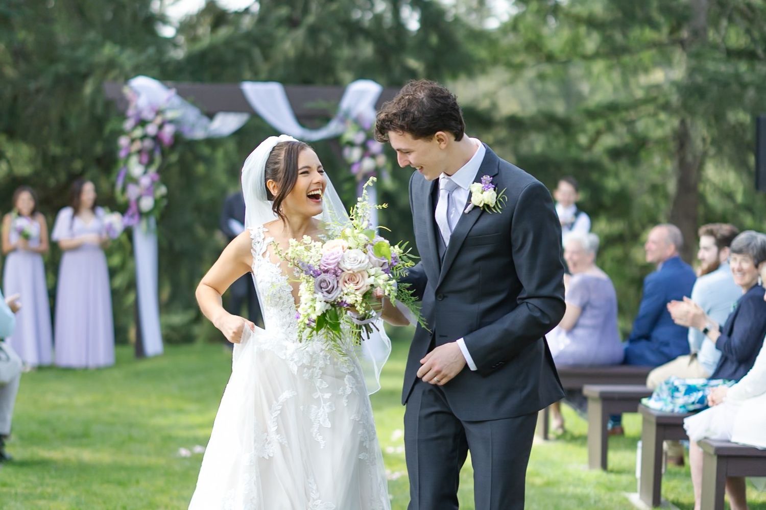Joyful bride and groom walking down outdoor wedding aisle, bride holding floral bouquet, bridesmaids in lavender dresses.