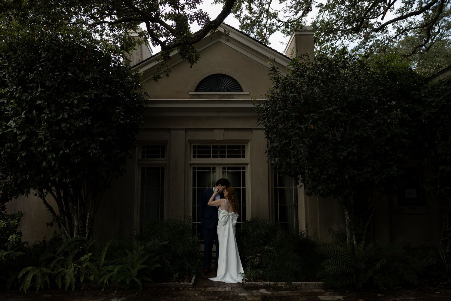 Couple embraces on a tree-lined path before an elegant Southern mansion during their wedding day.