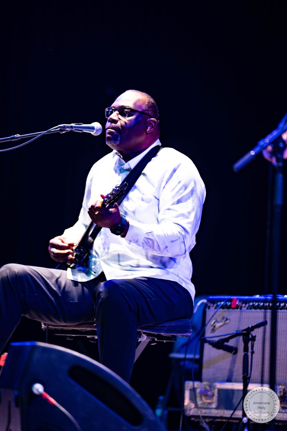 A Black male musician plays electric guitar and sings into a microphone on a dimly lit stage wearing a white shirt.