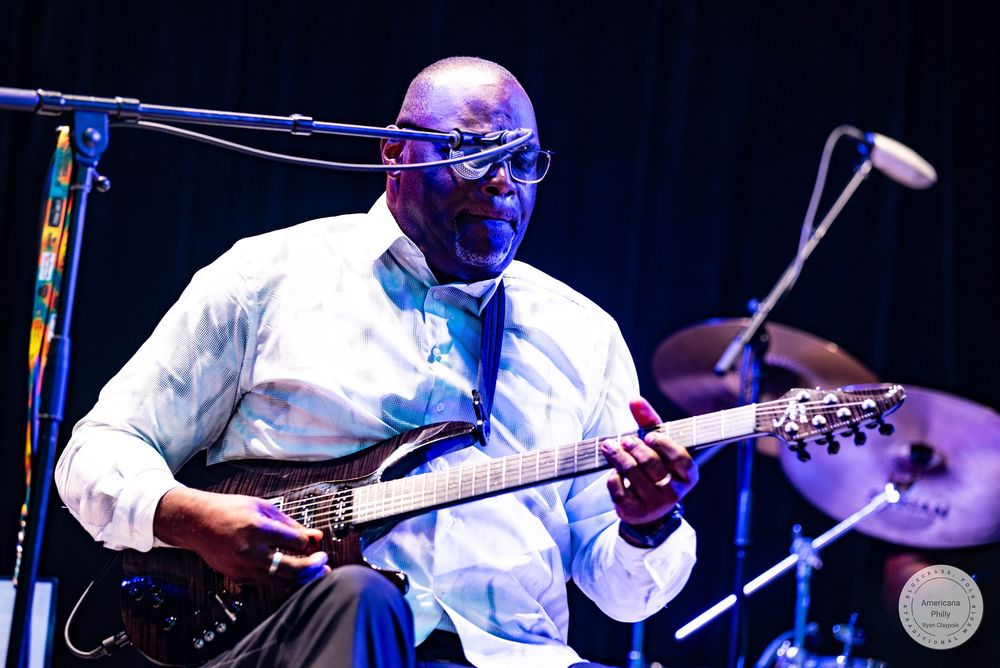 A musician wearing glasses and a white shirt plays electric guitar on stage under blue lighting.