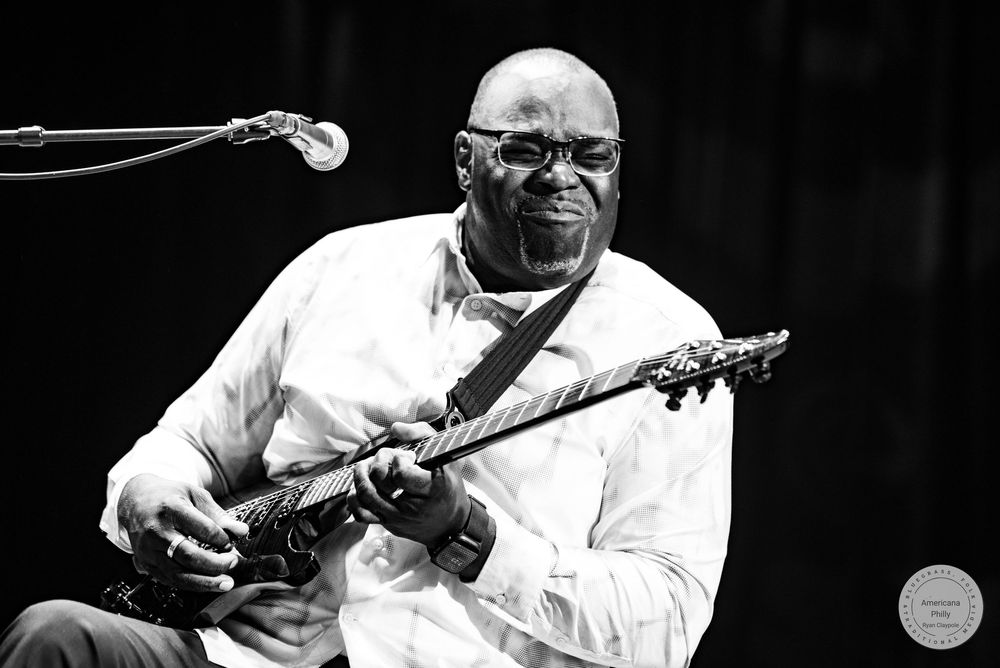 Black and white photo of a musician playing electric guitar on stage, smiling joyfully during a live performance.
