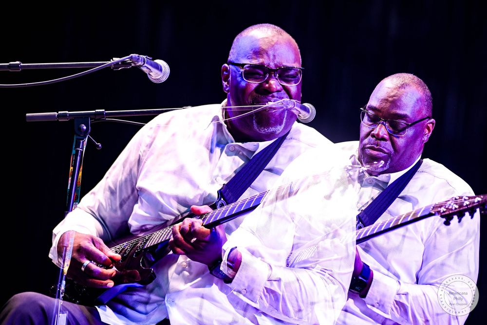 Two musicians in white shirts perform on electric guitars under stage lighting.