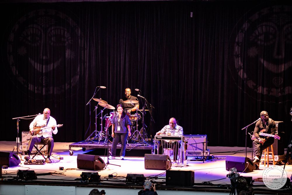 Live jazz band performing on stage with drummer, keyboardist, and guitarist under purple stage lights.