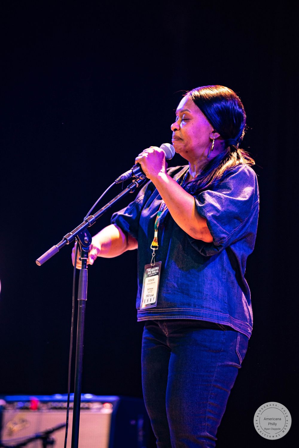 Woman singing into microphone on stage at live music event, wearing denim jacket with lanyard badge.