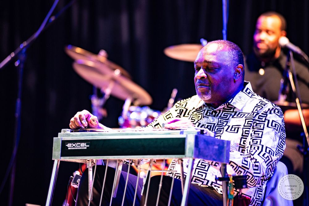 Musician playing a steel guitar on stage wearing a patterned shirt, with a drummer performing in the background.