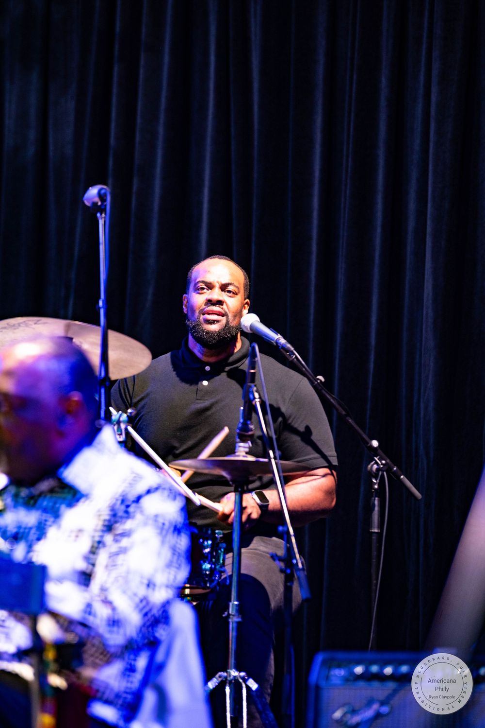 A Black male vocalist performs on stage at a live music event, singing into a microphone wearing a black polo shirt.