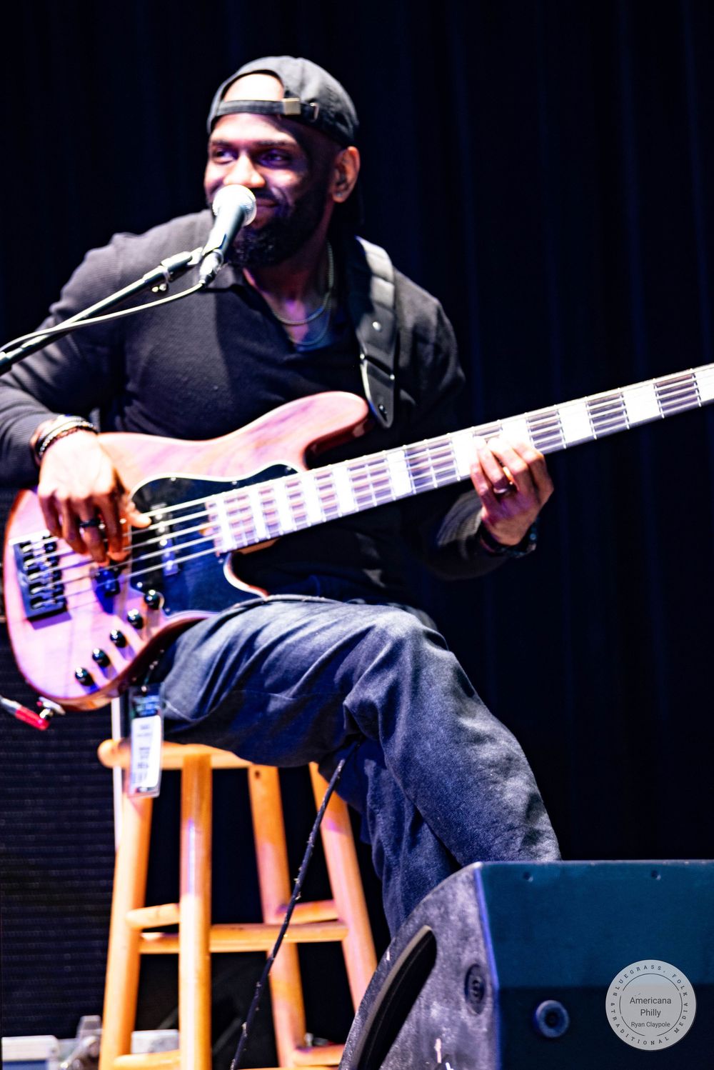 Musician playing bass guitar on stage, seated on stool at microphone during live performance.