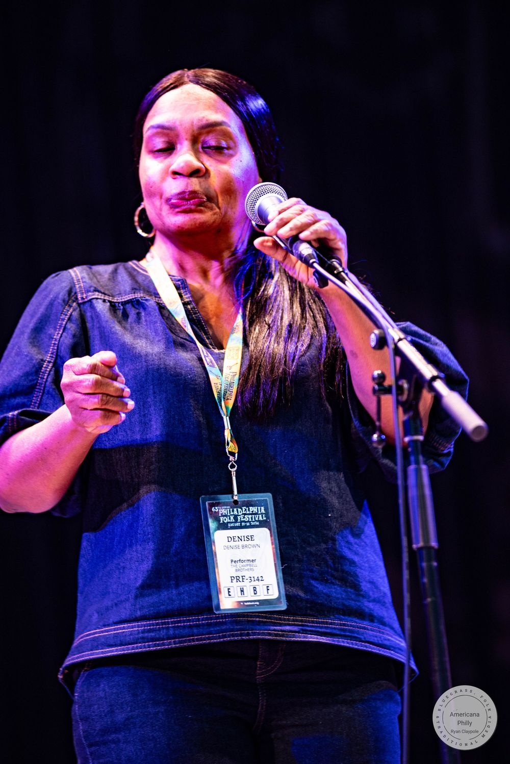 Woman named Denise singing into microphone on stage wearing denim outfit and lanyard badge at live music event.
