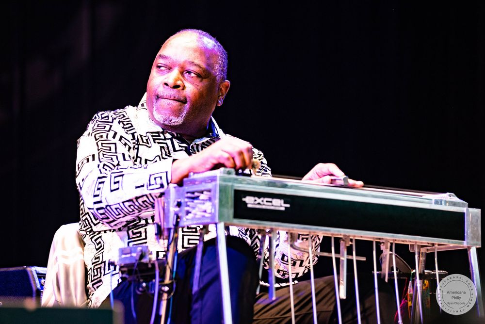 Man in patterned jacket playing a steel guitar on stage under bright lights.