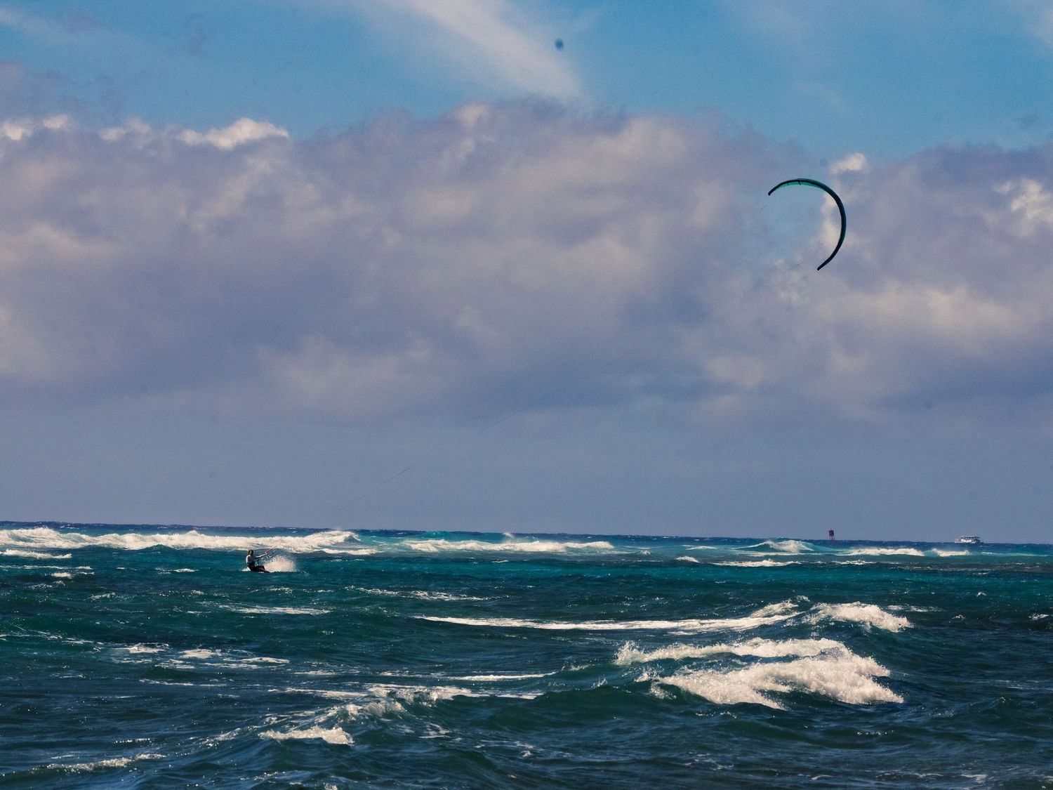 A kitesurfer skims across choppy blue-green water at Waikiki, a curved kite arcing high against a bright sky of scattered clouds.